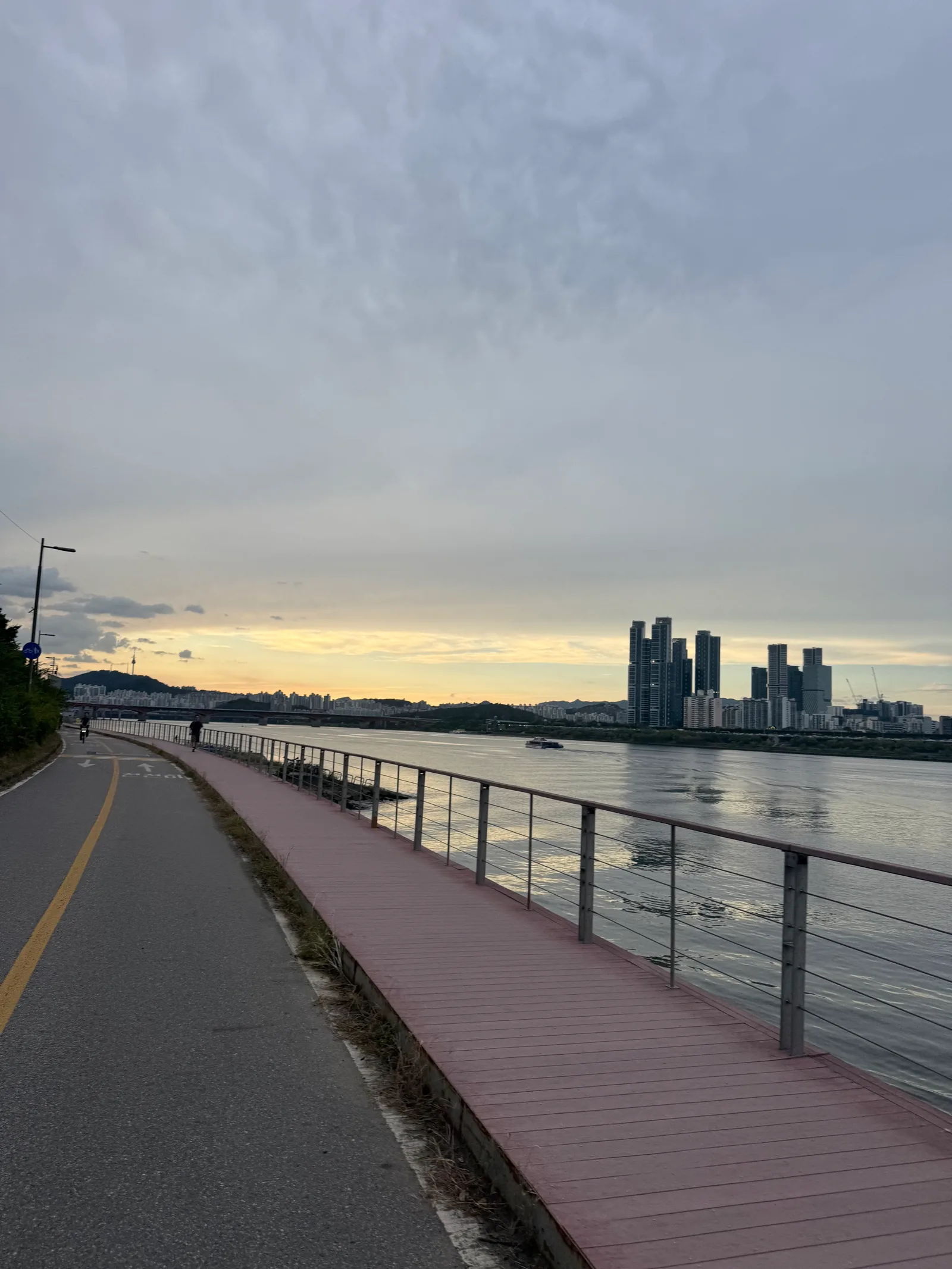 Riverside bike lane and red wooden boardwalk at dusk, apartments and a bridge across the water