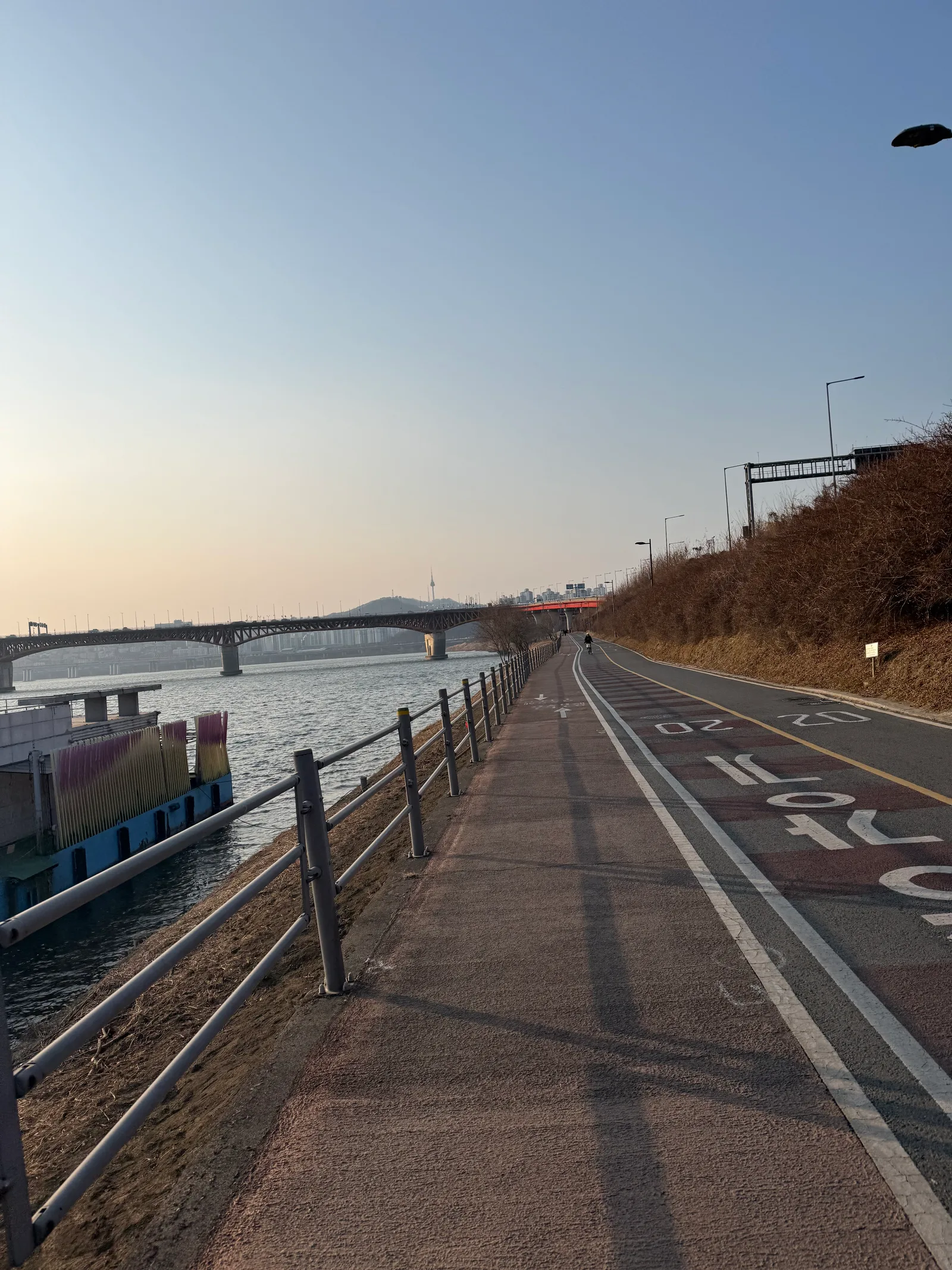 Han River bike lane beside a pedestrian path, with a 20 marking on the surface and a steel truss bridge over the river