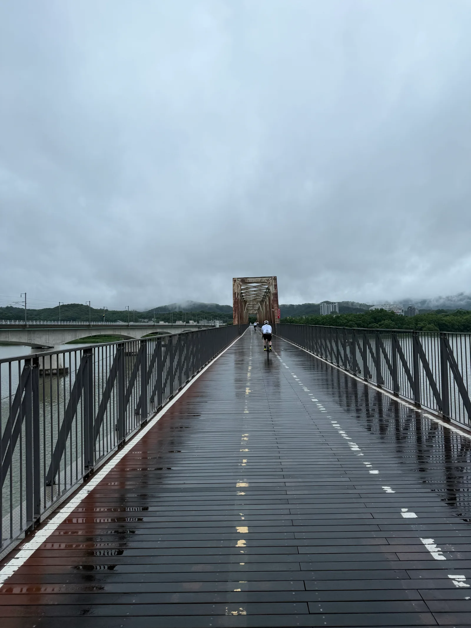 Two riders moving away along a wet bike-only bridge deck under a cloudy sky