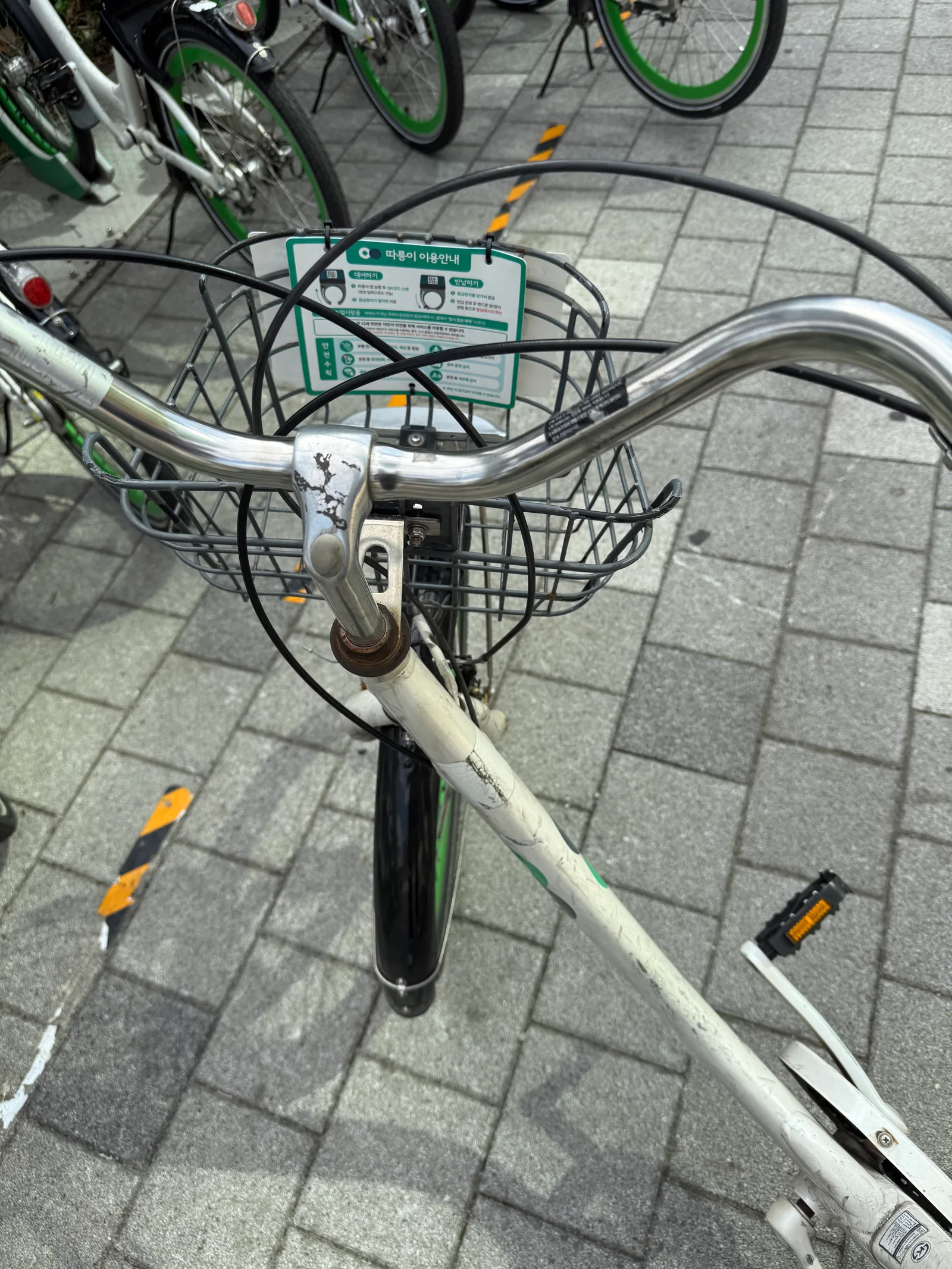 Handlebar and front basket of a white public bike, with a plate reading 따릉이 이용안내 / 대여하기 / 반납하기 / 안전수칙 / 임시잠금